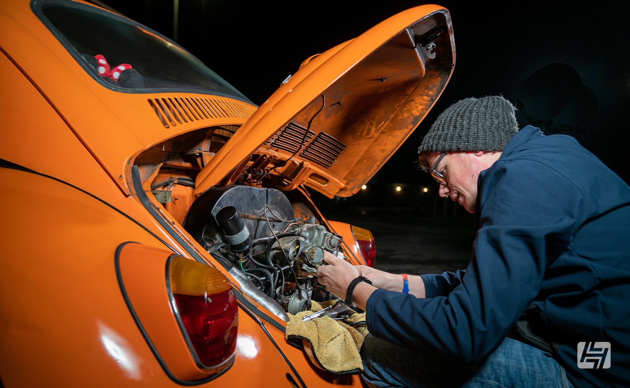 enthusiast removing carburettor from orange VW Beetle at night 