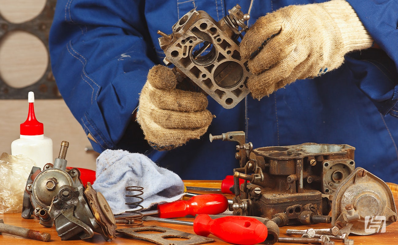 Mechanic in blue overalls and white gloves takes a carburettor apart on a workbench