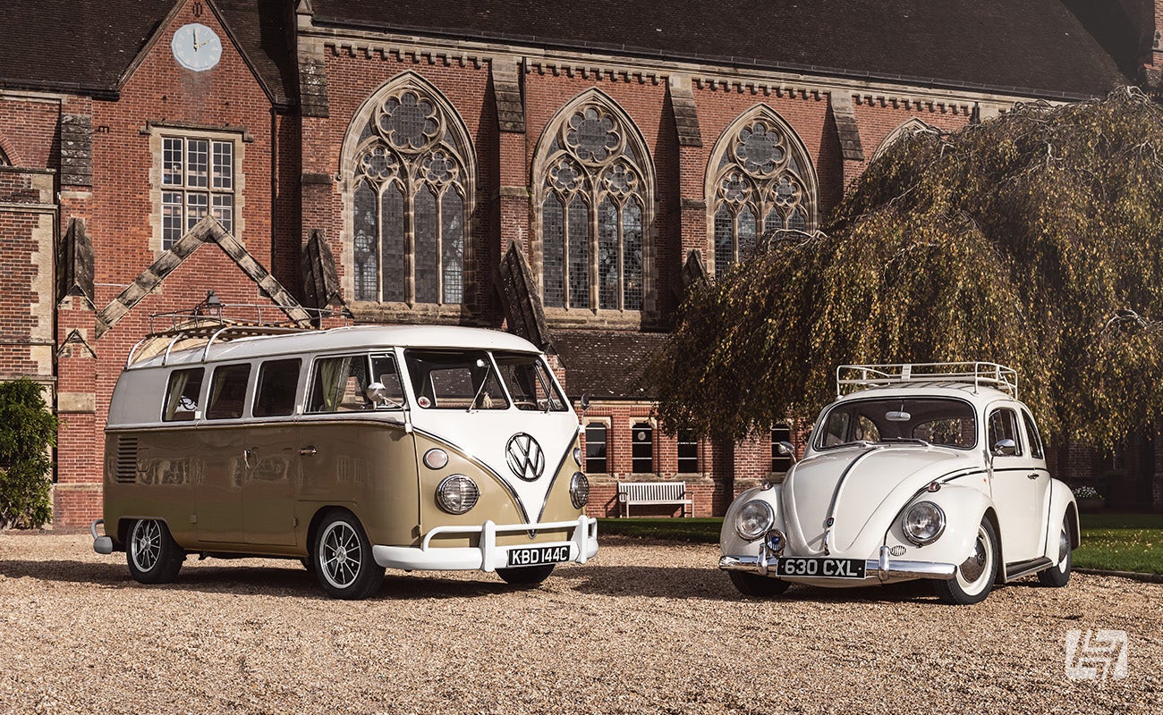 VW Split Bus and white VW Beetle parked together on gravel driveway